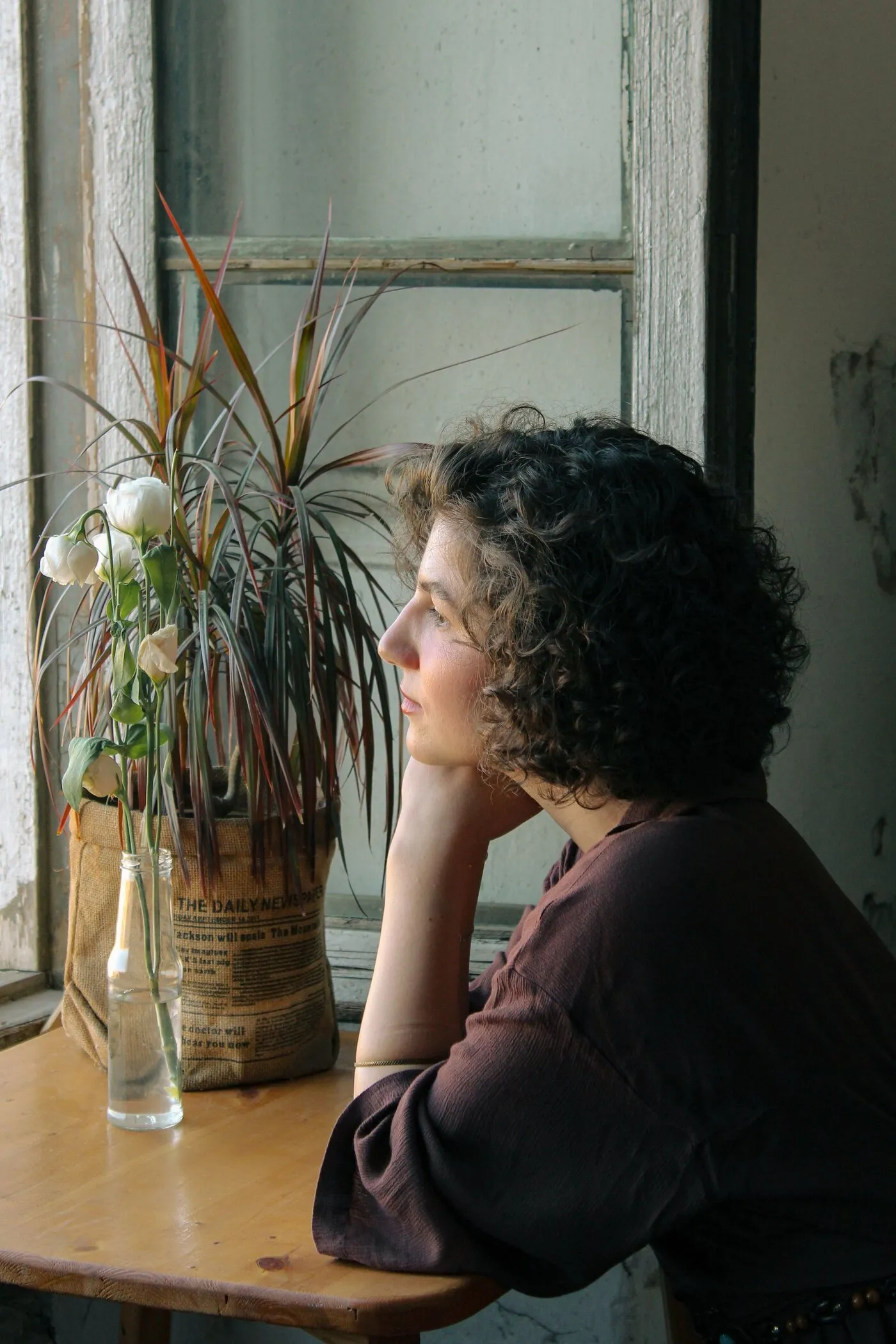 A woman rests her chin on her hand at a table by a window and looks outside. A plant and flowers stand beside her.