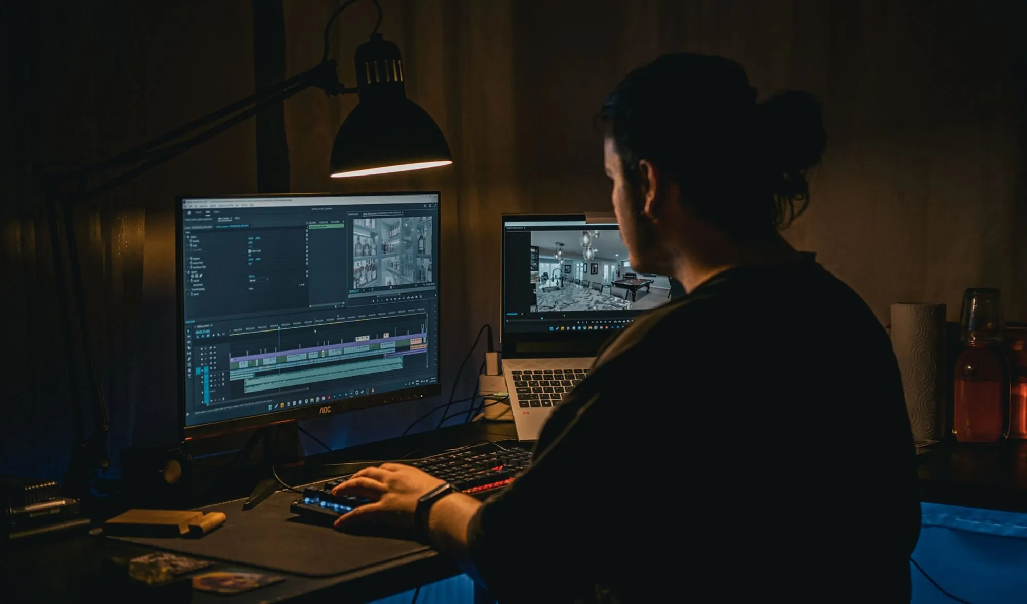 A video editor works at night under a desk lamp, with a timeline on the monitor and a laptop beside it.