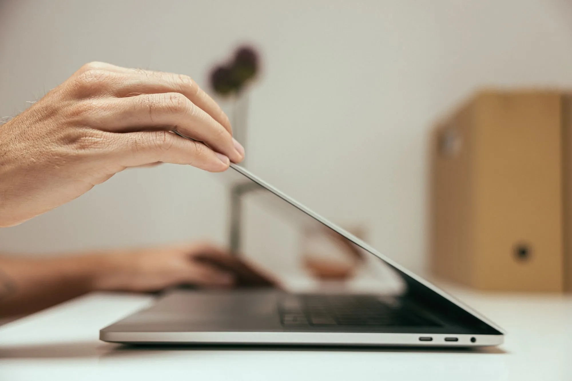 A hand lifts the lid of a laptop on a white desk.