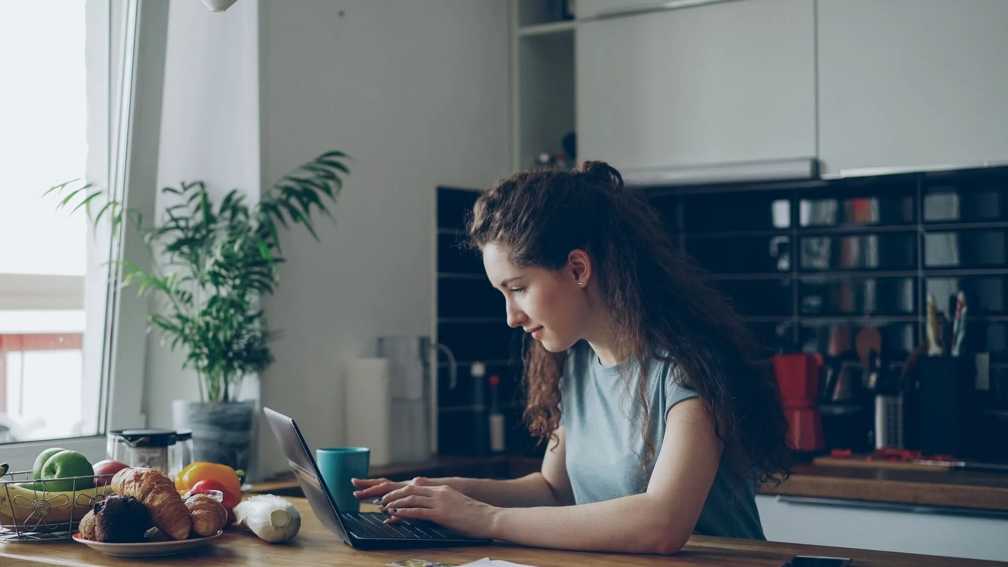 A woman types on a laptop at a kitchen table in the morning. Fruit, bread and a plant sit beside her.
