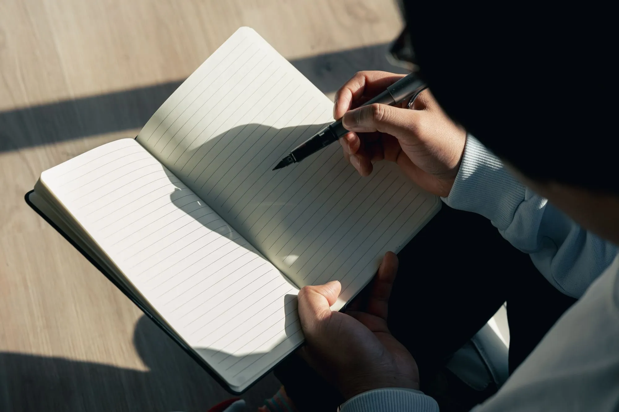 A hand holds a pen over the blank lined page of a notebook in sunlight.