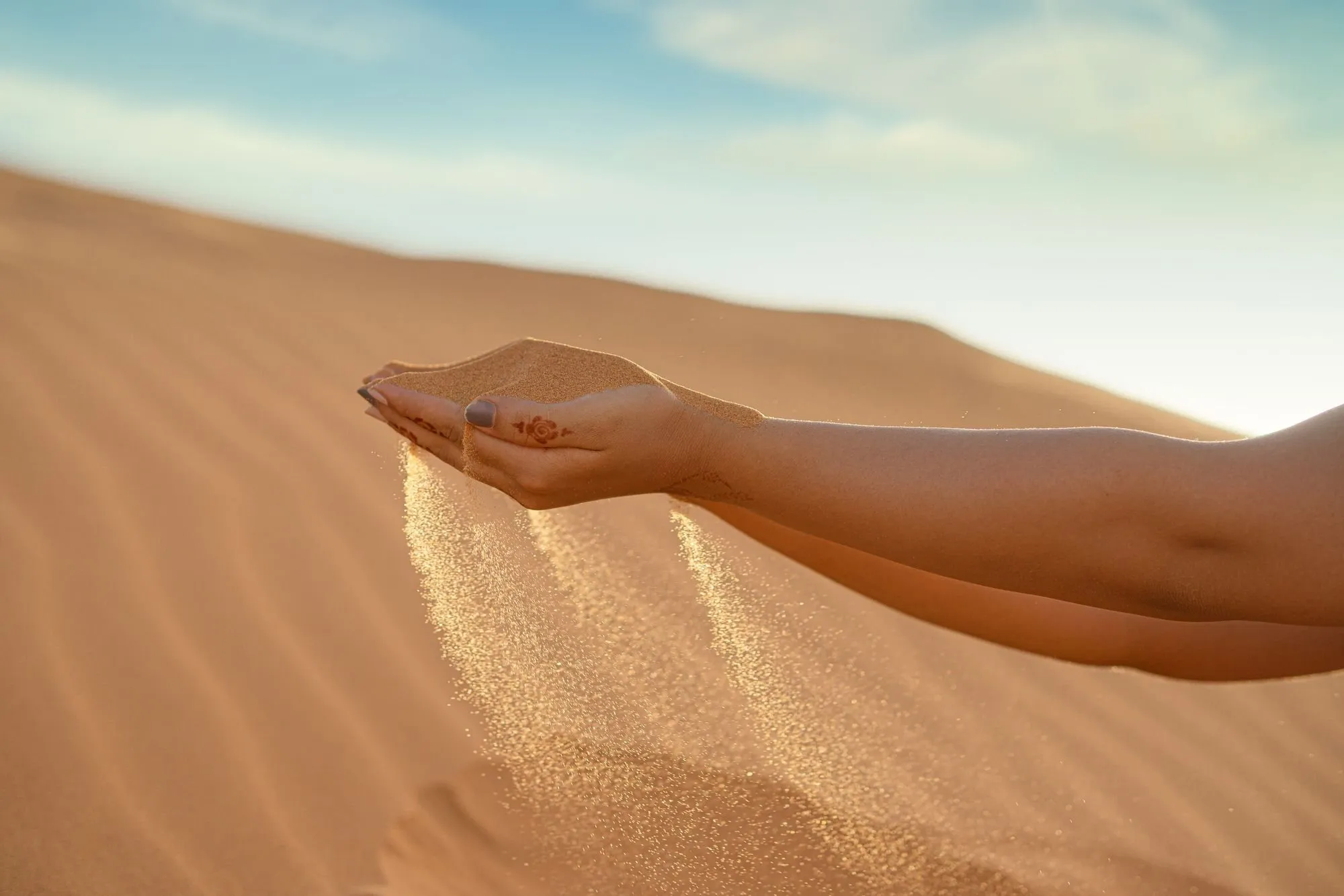 Fine sand pours from a cupped hand onto a desert dune in warm light.