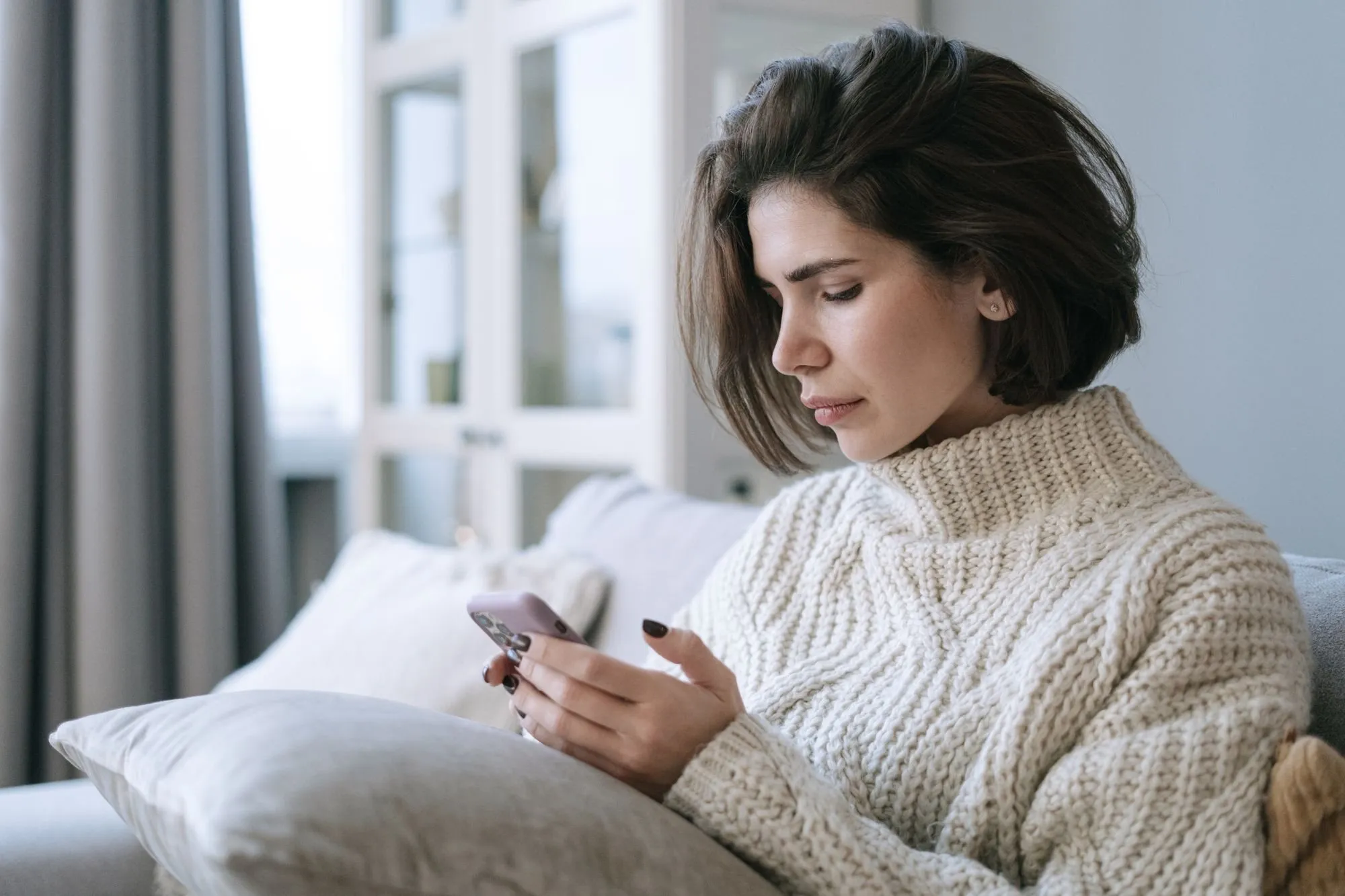 A woman in a cream knitted sweater sits on a sofa and looks down at her phone.
