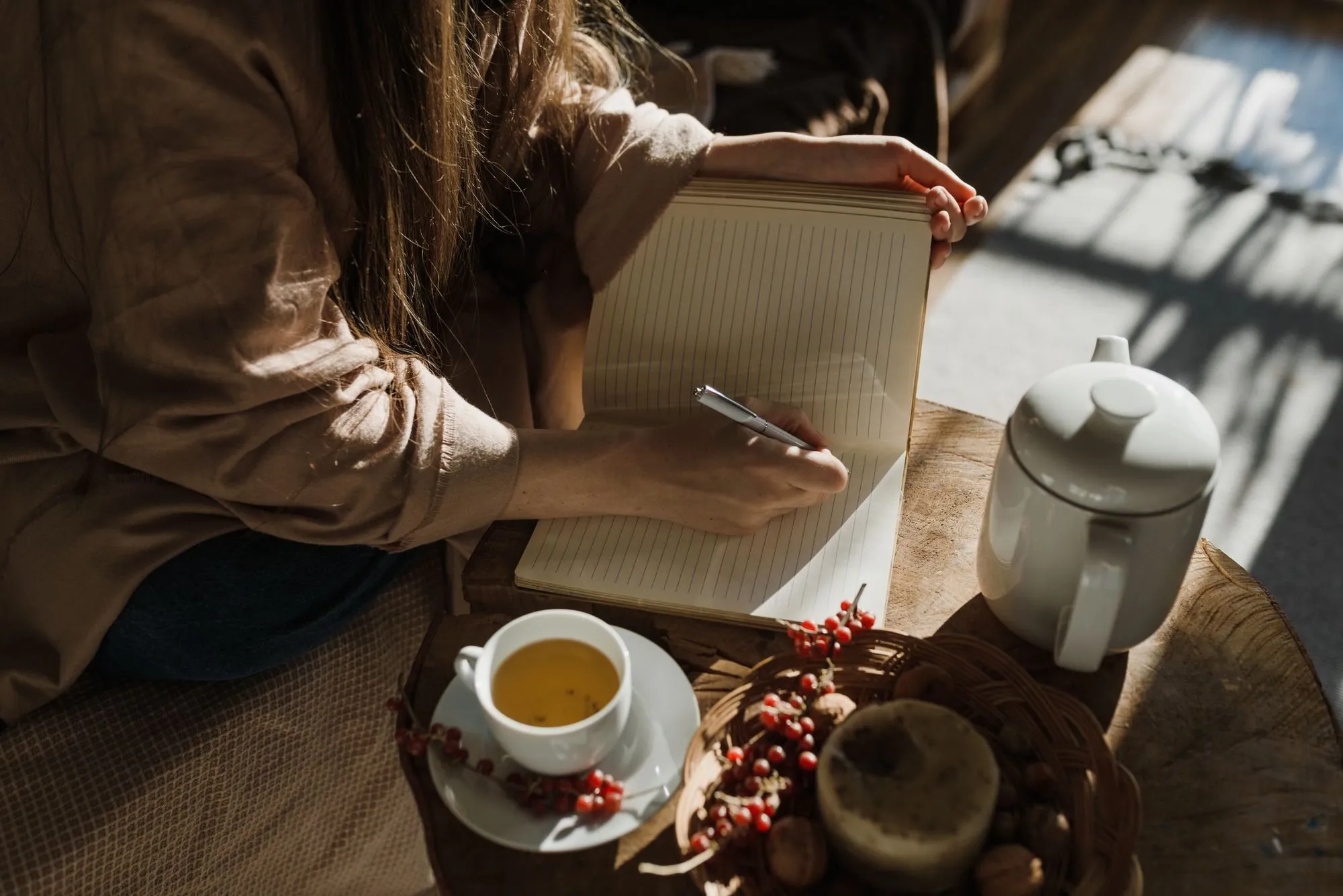 A woman writes in an open notebook in low sun, with a cup of tea and a white teapot beside her.